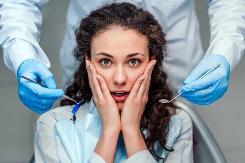 a young woman holds her face between her hands and looks anxious while a dentist stands behind her with dental instruments in their hands