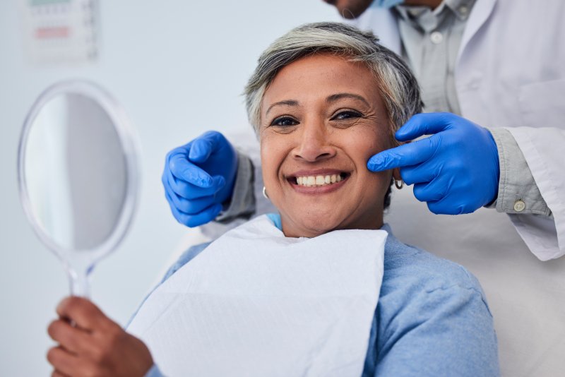 Older woman sitting in dental chair holding mirror up to her face while smiling as dentist points at her mouth behind her